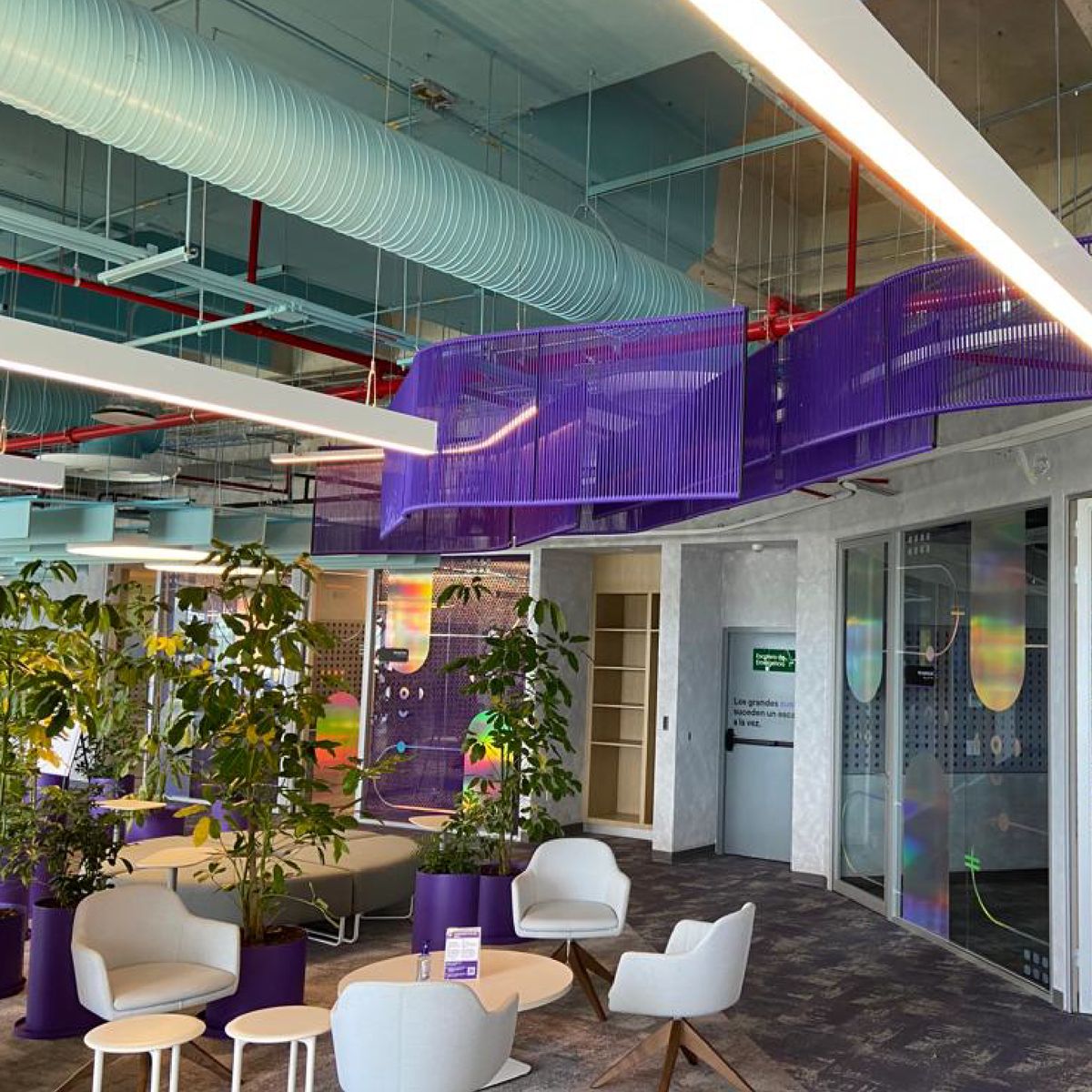 Plafón ondulado de cordón morado suspendido bajo ductos turquesa y tuberías rojas, sobre una sala de oficina con sillones blancos, mesas redondas y macetas moradas con plantas