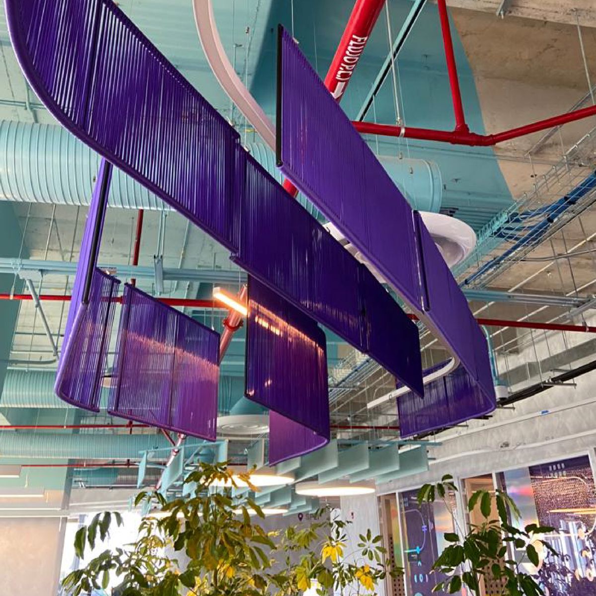 Paneles curvos de cordón morado colgados del techo, vistos desde abajo, contra ductos turquesa, tuberías rojas y la copa de unas plantas