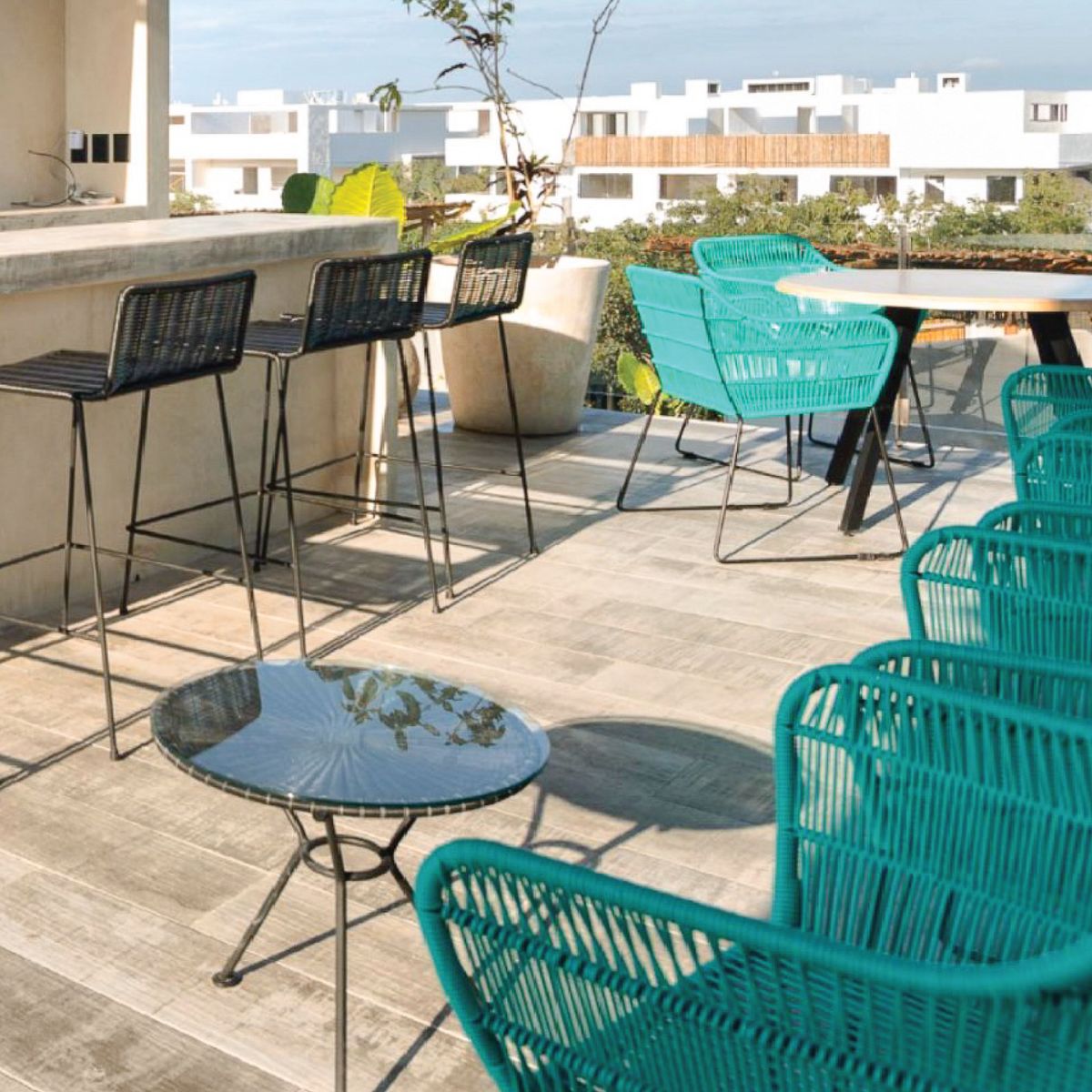 Terraza al sol con sillas de cordón turquesa en primer plano, bancos altos de tejido negro junto a una barra de concreto y una mesa baja de cristal