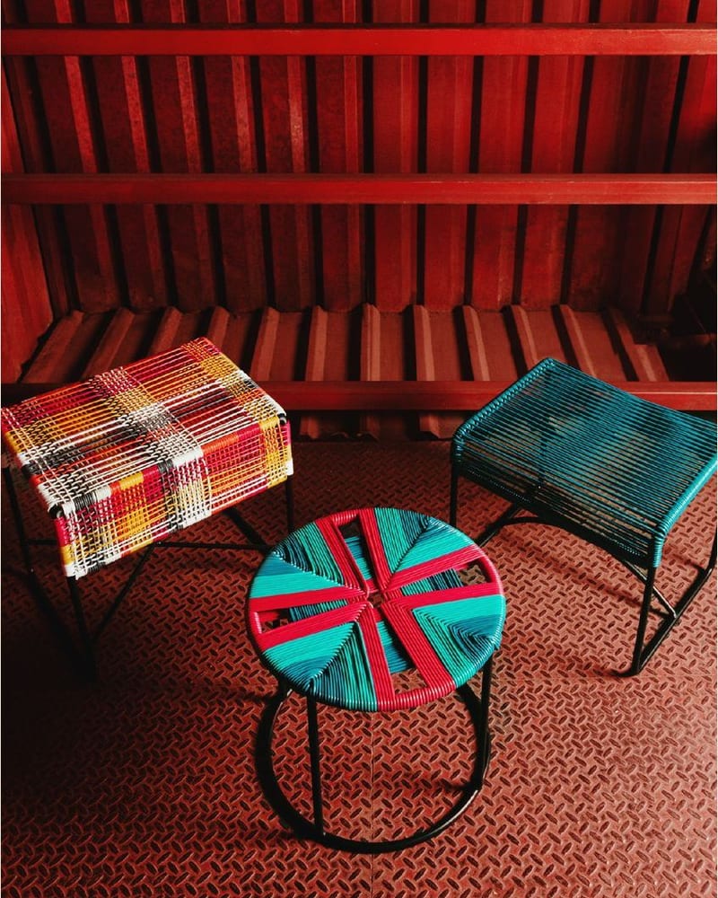 Dos bancos tejidos y una mesa redonda de tapa multicolor sobre un piso de lámina antiderrapante roja, bajo un techo de vigas del mismo rojo
