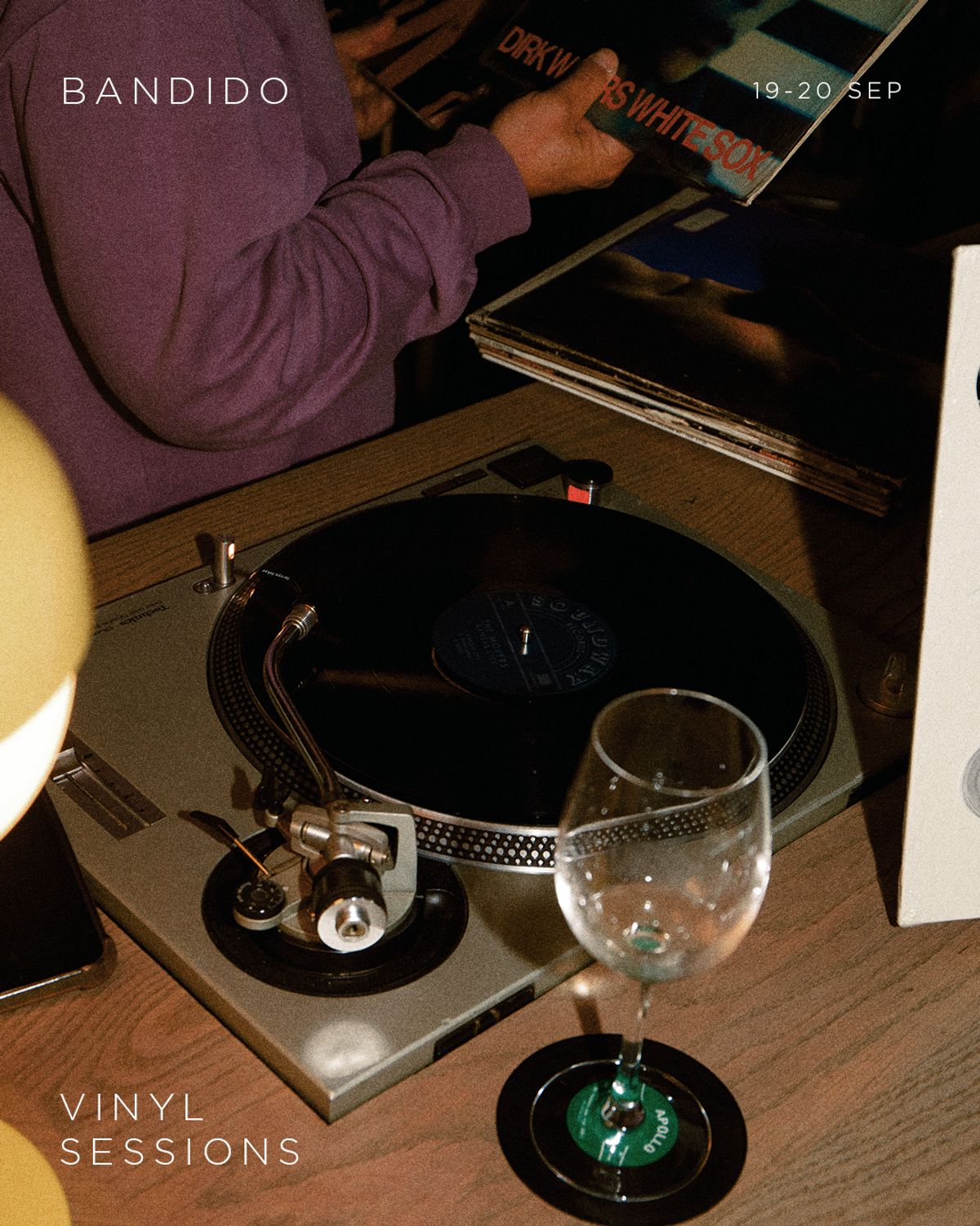Fotografía con luz baja de una tornamesa plateada con un disco girando y una copa vacía sobre un posavasos de vinilo; al fondo, unas manos en sudadera morada sostienen un LP, con el texto BANDIDO, 19-20 SEP y Vinyl Sessions en letra fina blanca