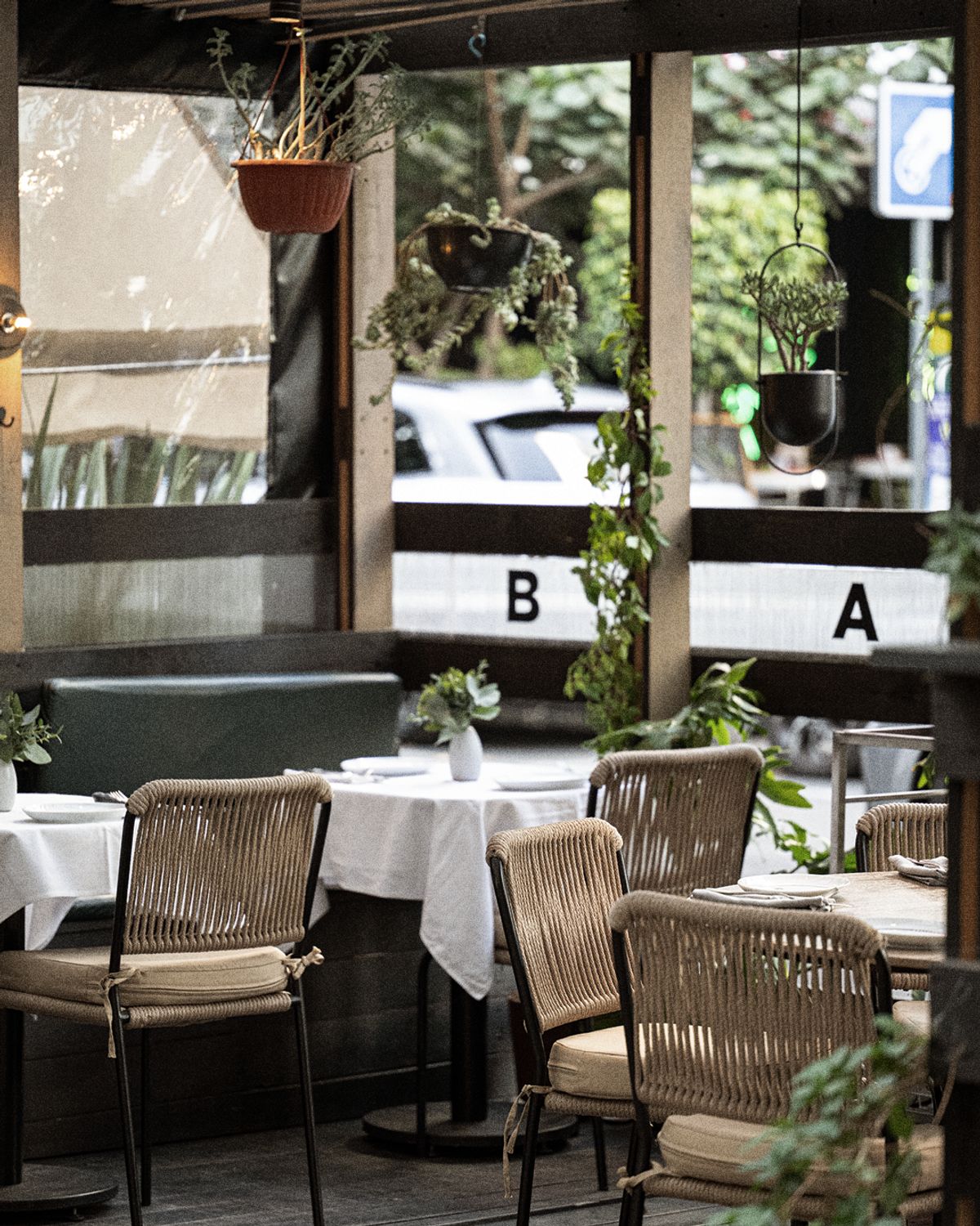 Terraza de Bandido sin gente: sillas de cuerda trenzada, mesas con mantel blanco y macetas colgantes, con las letras B y A de la marca rotuladas en negro sobre el barandal