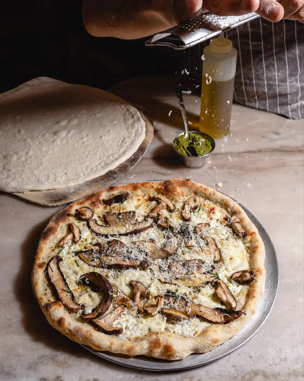 Una mano ralla queso sobre una pizza blanca de hongos recién salida del horno en Fabiana's, con luz natural y el borde tostado: el tipo de foto de producto que conviene subir al perfil