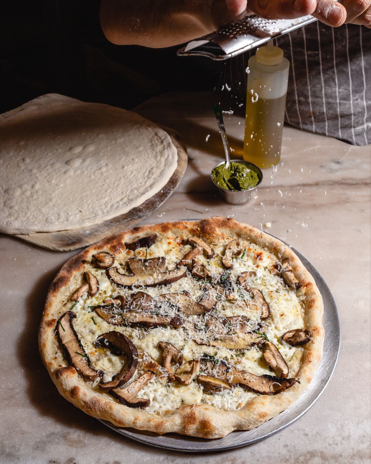 Una mano ralla queso sobre una pizza blanca de hongos recién horneada, con el borde tostado, sobre una barra de mármol junto a una masa extendida, un frasco de aceite y un tazón de pesto