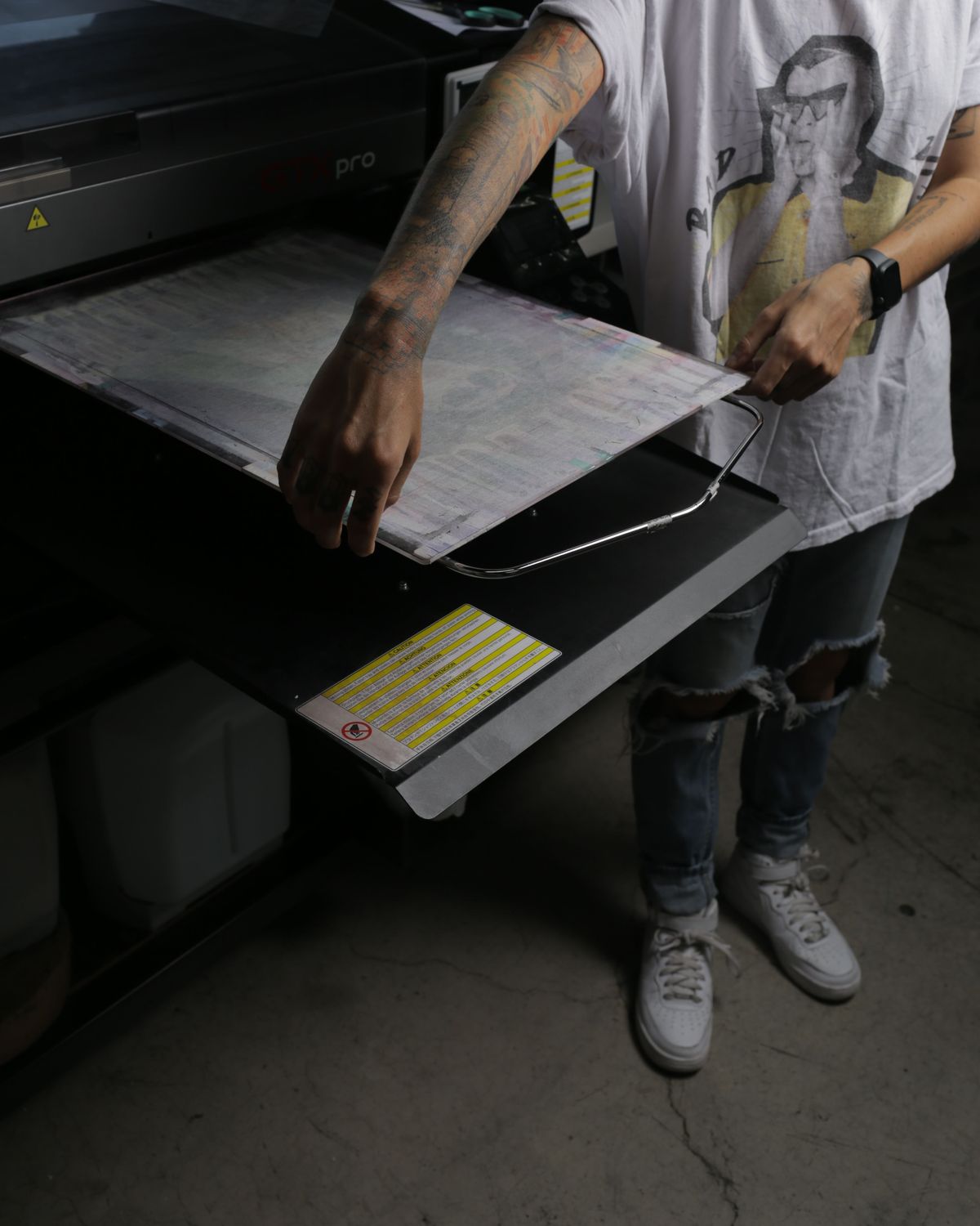 En el taller, una persona con los brazos tatuados desliza la base de una impresora textil GTX pro; se ve de los hombros para abajo, con playera estampada, jeans rotos y tenis blancos
