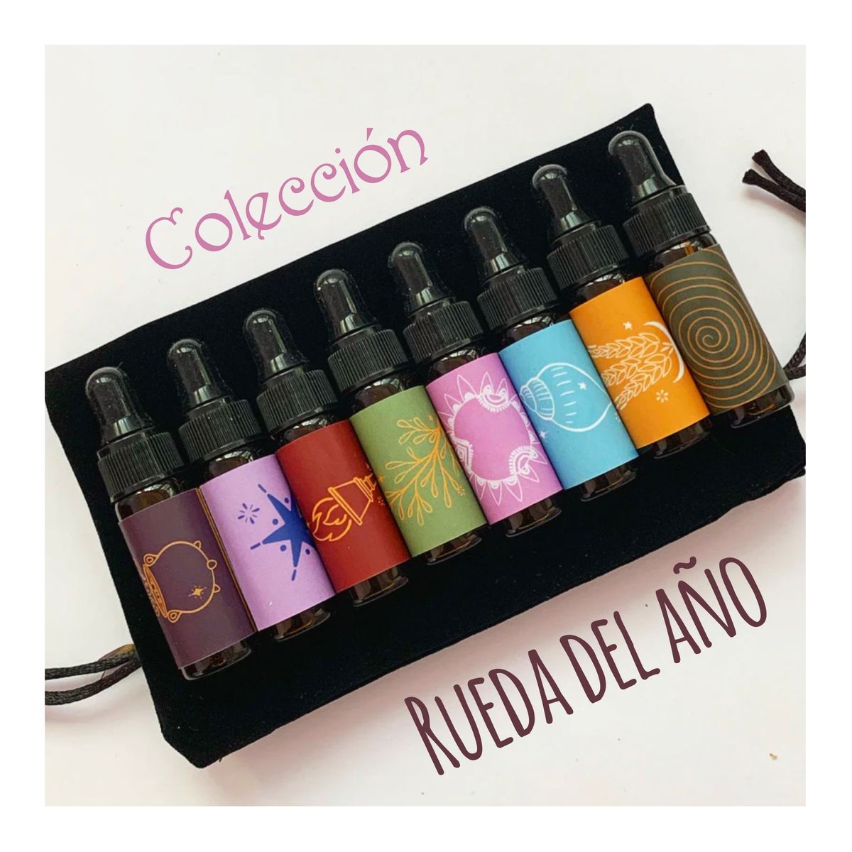 Ocho goteros de vidrio ámbar sobre un estuche de terciopelo negro, cada uno con una etiqueta de color ilustrada (caldero, estrella, campana, rama, corazón, caracol, espiga y espiral), con los textos «Colección» y «Rueda del año»