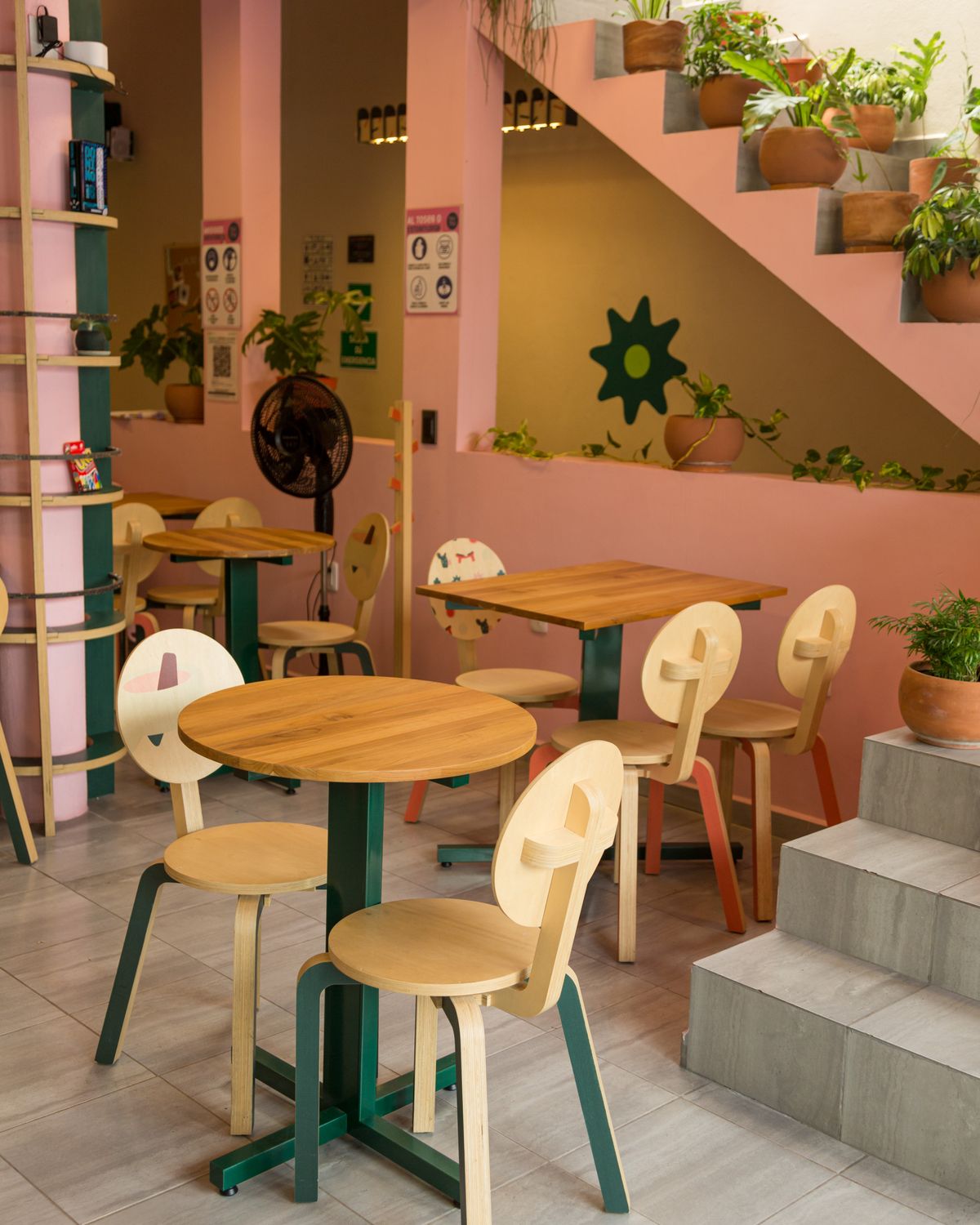 Salón con mesas redondas de madera y sillas con respaldo recortado, junto a una escalera de concreto con macetas y muros rosa y verde