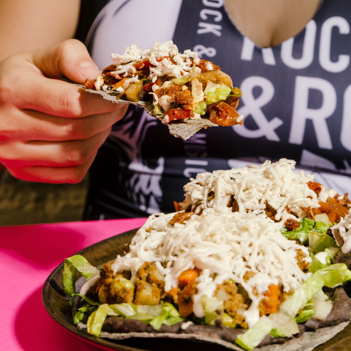 Una mano levanta un trozo de tostada de maíz azul con guisado rojo, lechuga y queso vegano rallado, sobre un plato de tostadas servido en una mesa rosa