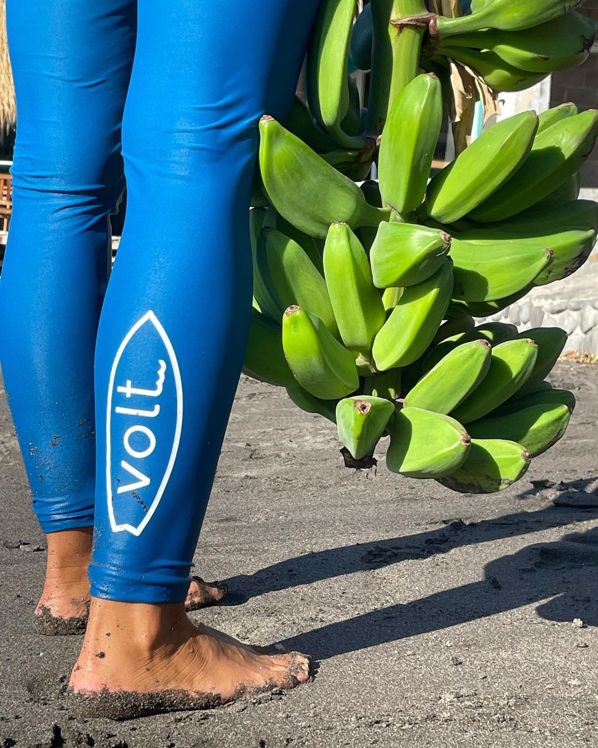 Detalle: las piernas en el traje azul con el logotipo «volt.» en blanco, los pies descalzos en la arena oscura y el racimo de plátanos colgando al lado