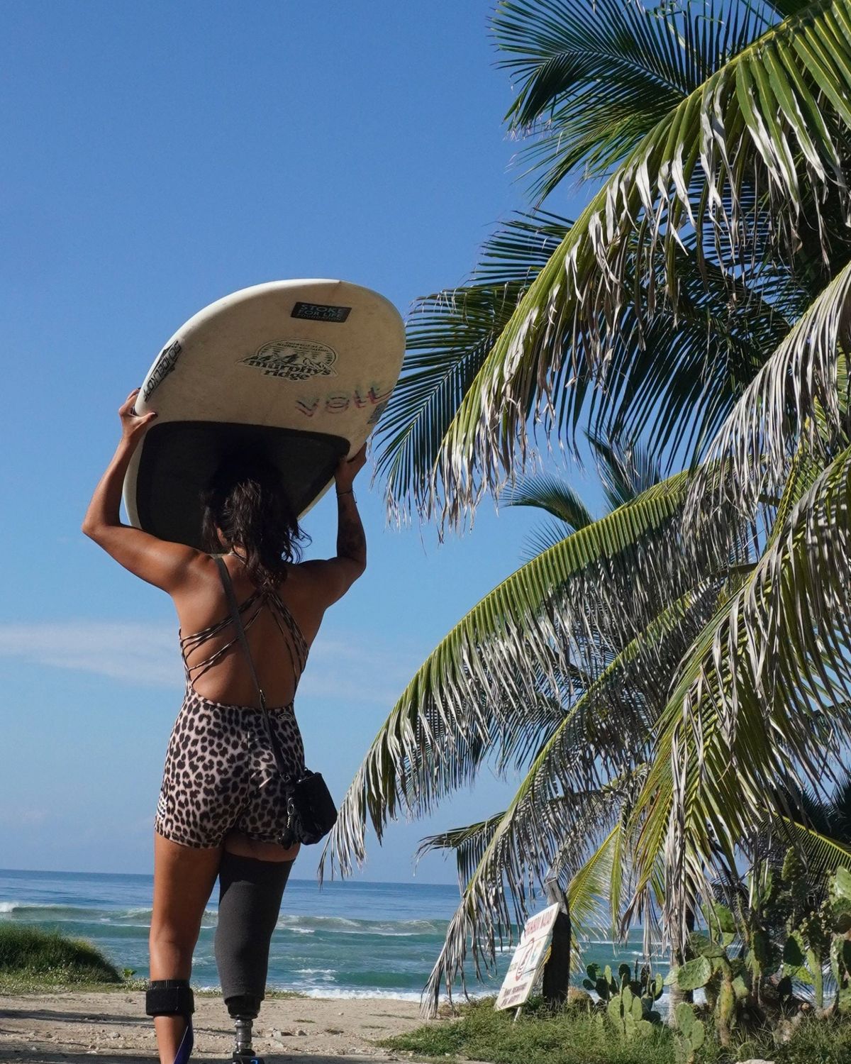Una surfista de espaldas, con traje estampado de leopardo, sostiene la tabla sobre la cabeza entre palmeras, con el mar al fondo