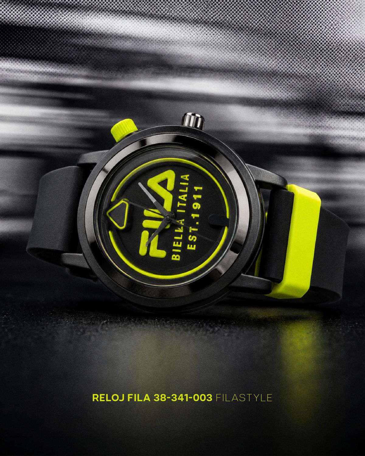 Reloj FILA negro con carátula y correa en amarillo neón, inclinado sobre una superficie oscura que lo refleja, ante un fondo gris barrido en semitono; abajo, «Reloj FILA 38-341-003 Filastyle»