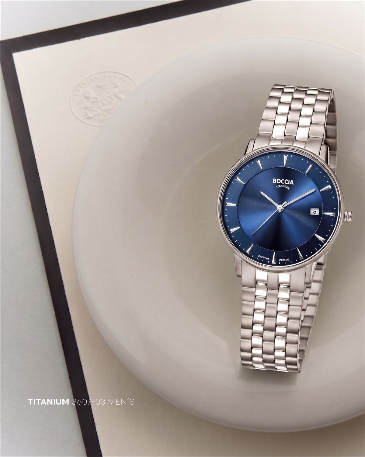 Reloj Boccia Titanium de acero con carátula azul solar, apoyado en un plato de cerámica blanca sobre papel crema con marco negro y un sello en relieve; abajo, «Titanium 3607-03 Men's»